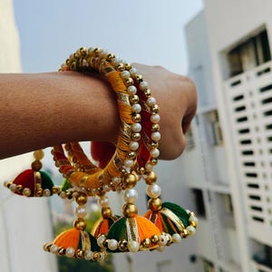 Pu&ograve; includere: Un set di tre braccialetti colorati con dettagli dorati e perline di perle bianche. Ogni braccialetto ha un fiocco che pende da esso, con colori arancio, verde e giallo.