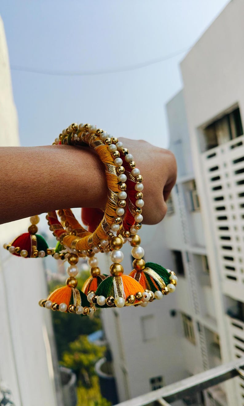 Pu&ograve; includere: Un set di tre braccialetti colorati con dettagli dorati e perline di perle bianche. Ogni braccialetto ha un fiocco che pende da esso, con colori arancio, verde e giallo.