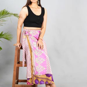 May include: A woman wearing a black sleeveless top and a pink and purple wrap skirt with a floral print. She is sitting on a wooden stool and smiling at the camera.