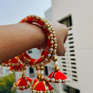 Pu&ograve; includere: Braccialetti rossi e dorati con accenti di perle bianche e nappe rosse e dorate. I braccialetti sono indossati al polso.