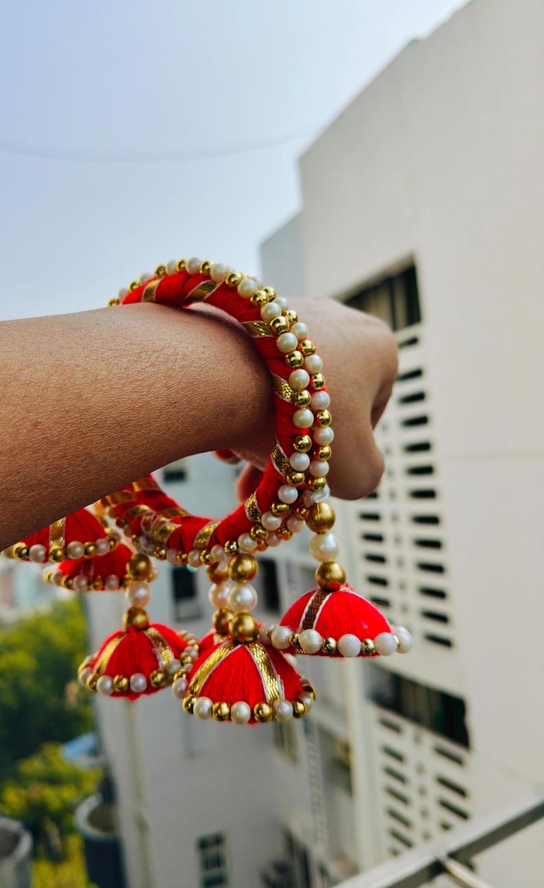 Pu&ograve; includere: Braccialetti rossi e dorati con accenti di perle bianche e nappe rosse e dorate. I braccialetti sono indossati al polso.