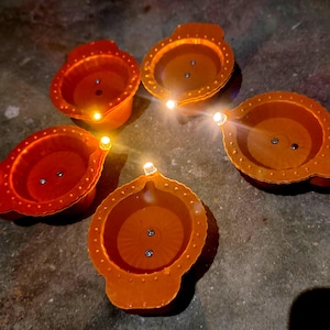 Puede incluir: Cinco lámparas diya de terracota naranja con un pequeño agujero en el centro de cada una. Las lámparas diya están iluminadas con pequeñas luces LED blancas.