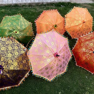 Pode incluir: Seis guarda-chuvas coloridos com padrões intrincados e guarnições de renda. Os guarda-chuvas estão dispostos em semicírculo sobre uma superfície de grama verde.