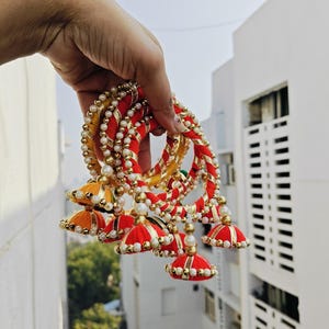 Pu&ograve; includere: Un set di braccialetti rossi e gialli con dettagli di perle bianche e finiture dorate. Ogni braccialetto ha un pendente a forma di nappa con una parte superiore in tessuto rosso e oro.