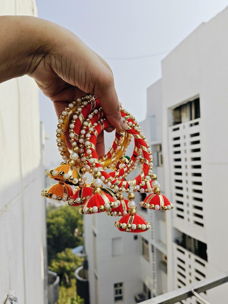 Pu&ograve; includere: Un set di braccialetti rossi e gialli con dettagli di perle bianche e finiture dorate. Ogni braccialetto ha un pendente a forma di nappa con una parte superiore in tessuto rosso e oro.