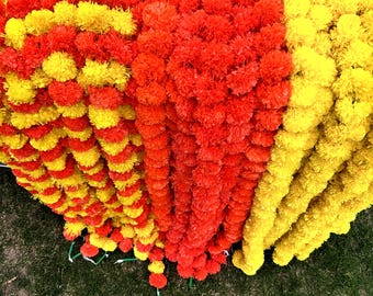 Può includere: Una ghirlanda di fiori di calendula artificiali in giallo e arancione. La ghirlanda è composta da più fili di fiori, creando un aspetto denso e pieno.