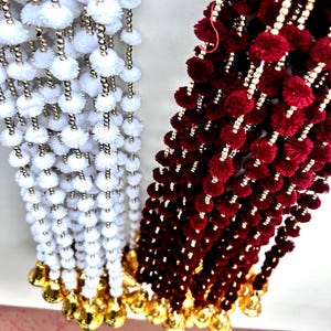 May include: Two strands of decorative trim with pom-poms. One strand is white with gold beads and the other is burgundy with gold beads.