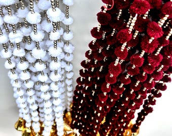 Può includere: Due fili di passamaneria decorativa con pompon. Un filo è bianco con perline dorate e l'altro è bordeaux con perline dorate.