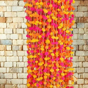 May include: A vibrant garland of colorful pom-poms in shades of pink, orange, and yellow, strung together with gold-colored beads. The garland is adorned with small, gold-colored bells at the bottom, creating a festive and decorative piece.