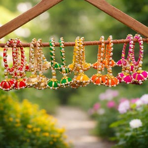 May include: A wooden hanger displays a collection of colorful, beaded ornaments. The ornaments feature red, gold, green, yellow, orange, and pink accents. Each ornament has a bell-shaped base and is adorned with pearls and beads.