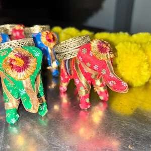 May include: Four small decorative elephant figurines made of fabric. The elephants are in red, green, and blue with gold accents. The elephants are sitting on a silver surface.