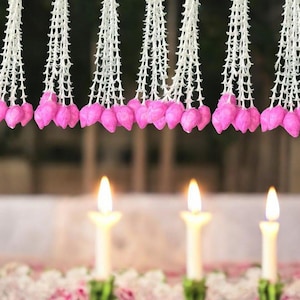 May include: Close-up of decorative garlands with white lace-like strands and pink flower-shaped ornaments. Three lit candles are in the foreground, with a blurred background. The scene evokes a sense of celebration or ceremony.
