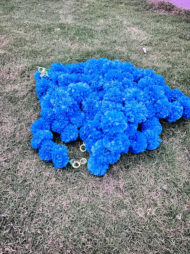 K&ouml;nnte beinhalten: Eine Girlande aus blauen k&uuml;nstlichen Blumen aus Papier, mit einem gr&uuml;nen Draht, der durch die Mitte verl&auml;uft. Die Girlanden liegen auf einem Bett aus gr&uuml;nem Gras.