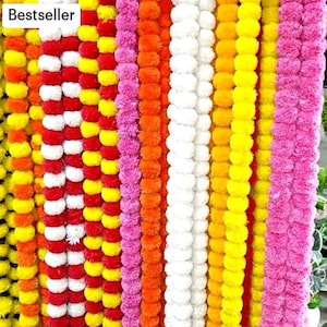 K&ouml;nnte beinhalten: Eine Sammlung bunter Girlanden aus Stoffpompons. Die Girlanden sind in den Farben Wei&szlig;, Rot, Gelb, Orange und Pink gehalten. Die Girlanden sind vertikal angeordnet und alle gleich lang. Der Text "Bestseller" ist in der oberen linken Ecke des Bildes sichtbar.