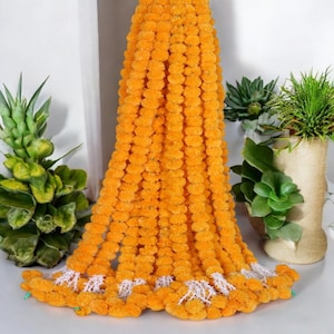 Könnte beinhalten: Eine lange Girlande aus leuchtend orangefarbenen Ringelblumen, mit einem weißen, spitzenartigen Besatz am unteren Ende. Die Blüten sind dicht gepackt und erzeugen ein volles, strukturiertes Aussehen. Grüne Pflanzen und eine dekorative Vase befinden sich im Hintergrund.