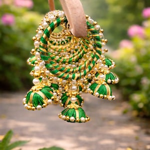 May include: Close-up of ornate green and gold earrings with pearl accents. The earrings are made of woven green and gold threads, with small pearl beads and bell-shaped dangles. The background is blurred, with greenery and soft lighting.