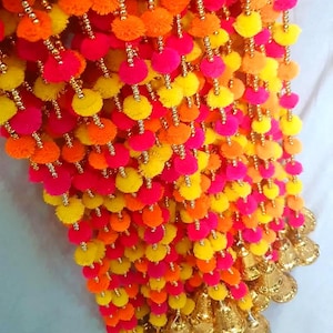 Könnte beinhalten: Eine lange Girlande aus bunten Pompons in Rosa, Gelb und Orange. Die Girlande ist mit goldenen Perlen besetzt und hat unten goldene Glöckchen.