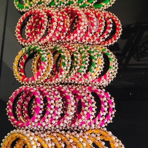 Può includere: Un mucchio di braccialetti colorati con accenti dorati e perline di perle bianche. I braccialetti sono fatti di tessuto e sono disposti in file di verde, rosa e giallo.