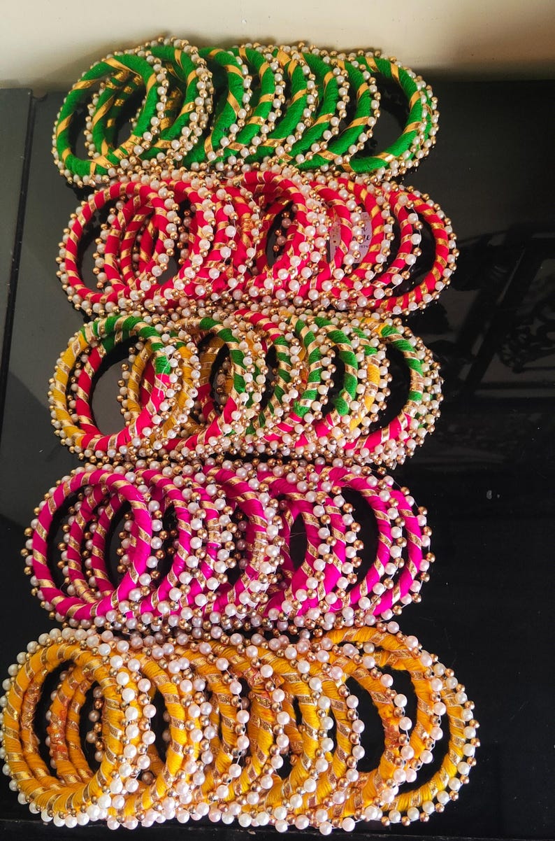 Pu&ograve; includere: Un mucchio di braccialetti colorati con accenti dorati e perline di perle bianche. I braccialetti sono fatti di tessuto e sono disposti in file di verde, rosa e giallo.