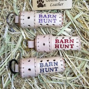 May include: Three white leather keychains with the words "BARN HUNT" in different colored lettering. The keychains are on a bed of straw. A small wooden sign with the words "Double Dew Leather" is in the background.