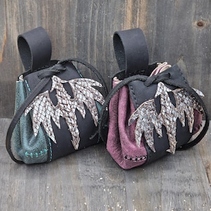 These small leather pouches feature a black drawstring closure and a hand-sewn silver dragon on the front. Each pouch includes a single belt loop and it is available in two colors: red or green.