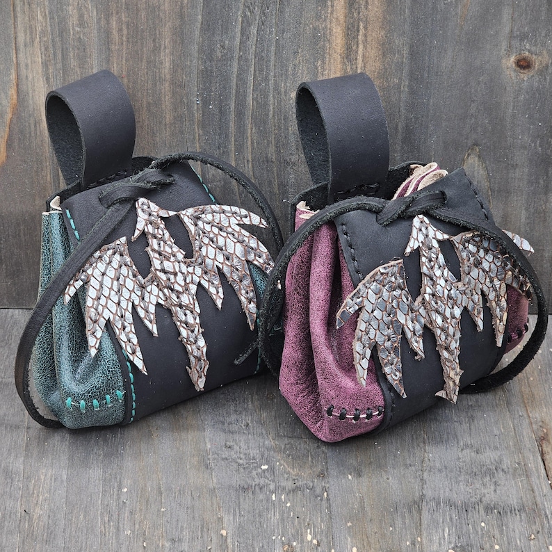 These small leather pouches feature a black drawstring closure and a hand-sewn silver dragon on the front. Each pouch includes a single belt loop and it is available in two colors: red or green.