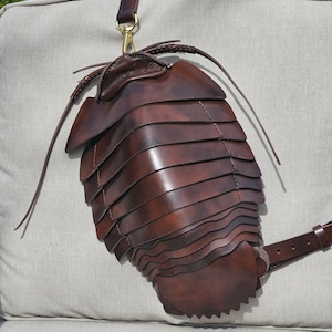 May include: A brown leather bag shaped like a segmented arthropod, such as a millipede or a pill bug. The bag has a strap and a buckle.