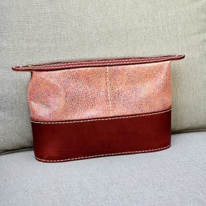 May include: A pink and red leather zippered pouch with a textured surface. The pouch has a white stitch detail around the edges.