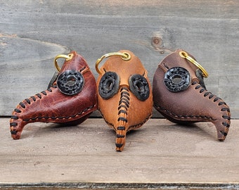 May include: Three leather keychains shaped like plague doctor masks. The masks are brown, tan, and dark brown with black eye holes. The keychains are hanging from gold rings.