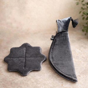 Puede incluir: Una bolsa para botella de vino de terciopelo gris oscuro con un posavasos a juego. La bolsa tiene una capucha con borla y botones decorativos. El posavasos tiene forma de trébol de cuatro hojas.