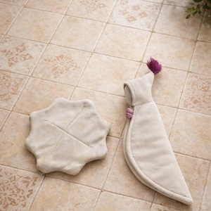 May include: Two beige suede items on a tiled surface. One is a star-shaped coaster with stitched details. The other is a curved, hooded item with a purple pom-pom and pink embellishments, possibly a bottle cover or warmer.