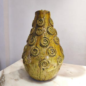 Puede incluir: Un jarrón de cerámica amarillo-verdoso hecho a mano con una superficie texturizada. El jarrón presenta una serie de diseños en espiral en relieve. El jarrón tiene una base bulbosa y se estrecha hacia la parte superior. El jarrón se exhibe sobre una superficie de mármol.