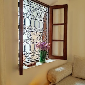 May include: A window with a decorative iron grate and a wooden frame. The window is open and there are purple flowers in a green vase on the windowsill.