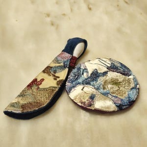 Puede incluir: Un conjunto de dos artículos de tela: una funda curva y acolchada y un posavasos redondo. La funda es azul marino con un estampado en beige, rojo y verde. El posavasos presenta un patrón similar en azul, blanco y morado.