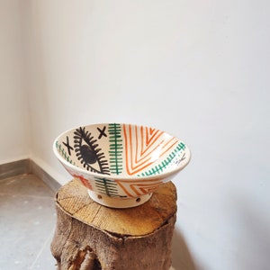 May include: A white ceramic bowl with a green, orange, and black geometric design. The bowl has a stylized eye with an X over it.