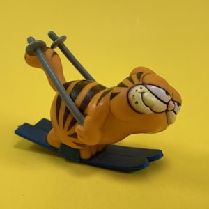 May include: A figurine of Garfield the cat skiing. The orange cat with black stripes is on blue skis, holding gray ski poles. The background is a solid yellow.