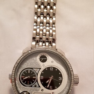 May include: A silver Techno Pave watch with a silver metal band. The watch face has a black dial with silver hands and a white dial with a silver hand. The watch face is surrounded by a row of small diamonds.
