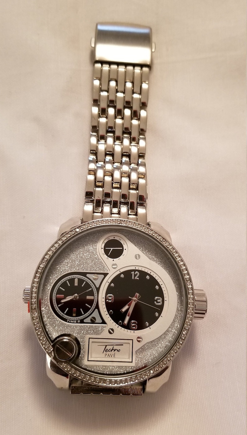 May include: A silver Techno Pave watch with a silver metal band. The watch face has a black dial with silver hands and a white dial with a silver hand. The watch face is surrounded by a row of small diamonds.
