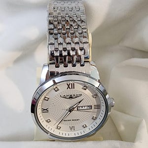 Op de afbeelding: Zilverkleurig polshorloge met een lichtgekleurde wijzerplaat en een schakelarmband. De wijzerplaat toont Romeinse cijfers, een datumvenster en de merknaam "LANMSOM". Het horloge is waterbestendig.