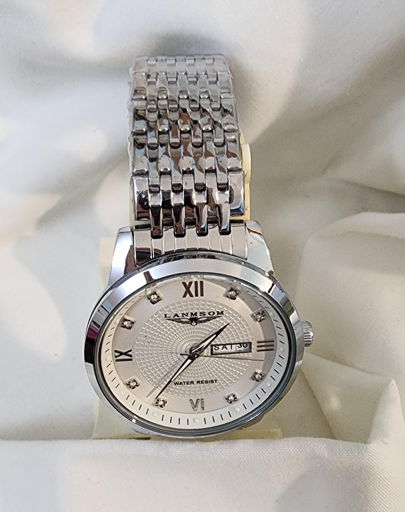 Op de afbeelding: Zilverkleurig polshorloge met een lichtgekleurde wijzerplaat en een schakelarmband. De wijzerplaat toont Romeinse cijfers, een datumvenster en de merknaam "LANMSOM". Het horloge is waterbestendig.