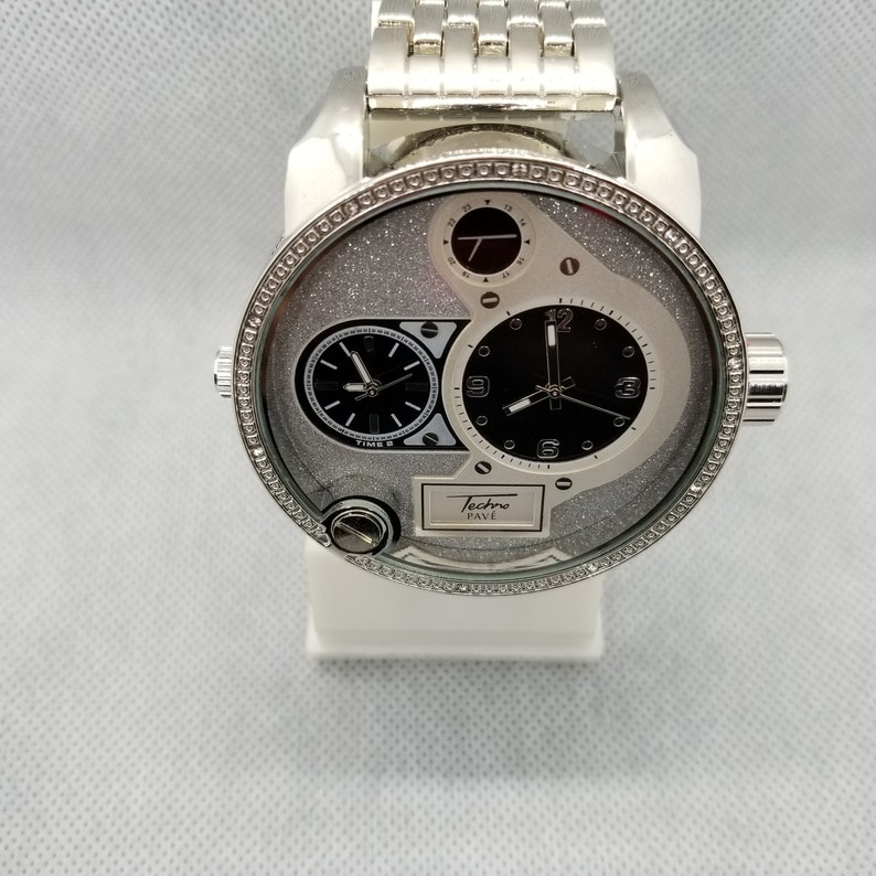 May include: A silver Techno Pave watch with a silver band and a white face with black hands and a silver glitter effect. The watch has a round face with a small round dial at the top and a larger round dial at the bottom.