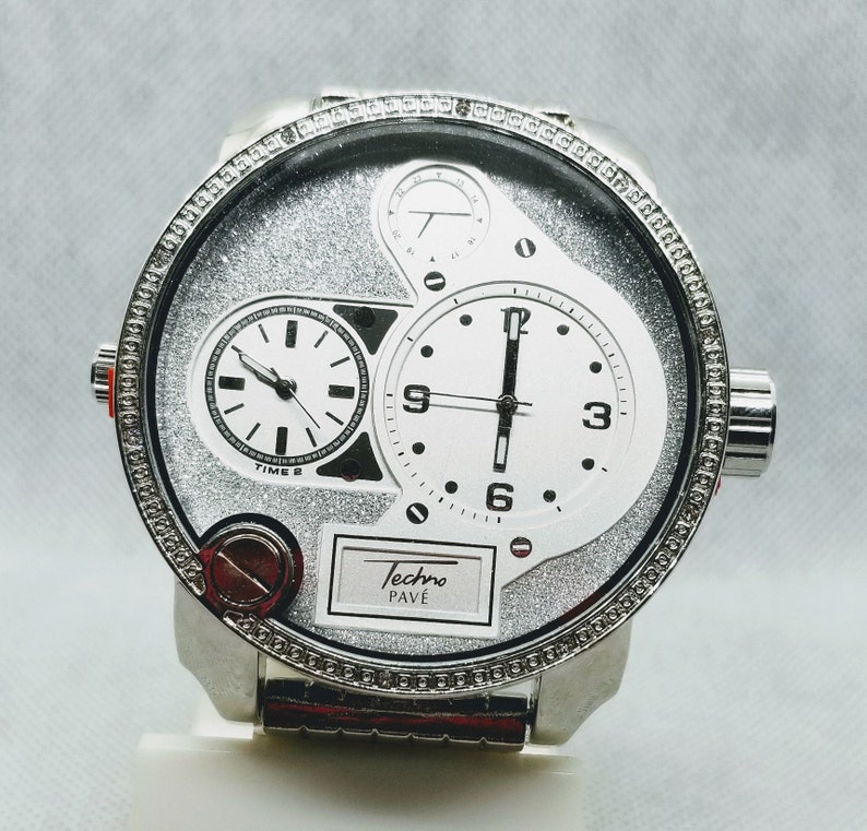 May include: A silver Techno Pav&eacute; watch with a silver band and a white face. The watch has three sub-dials, two of which are round and one of which is rectangular. The watch face is covered in a sparkly silver material.