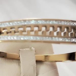 May include: A gold-toned bracelet with a zipper design. The bracelet features a row of clear crystals and a geometric pattern. The clasp is designed to look like a zipper pull. The bracelet is displayed on a white background.