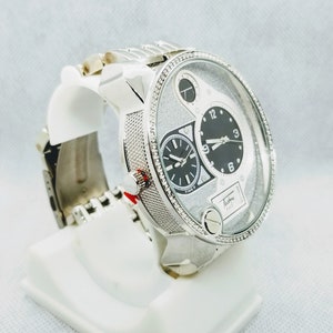 May include: A silver watch with a white face and black hands. The watch has a diamond-encrusted bezel and a silver band. The watch face has a small sub-dial at the 3 o'clock position and a larger sub-dial at the 9 o'clock position. The watch face also has the text "Techno Pave" printed on it.
