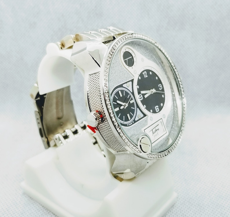 May include: A silver watch with a white face and black hands. The watch has a diamond-encrusted bezel and a silver band. The watch face has a small sub-dial at the 3 o'clock position and a larger sub-dial at the 9 o'clock position. The watch face also has the text "Techno Pave" printed on it.