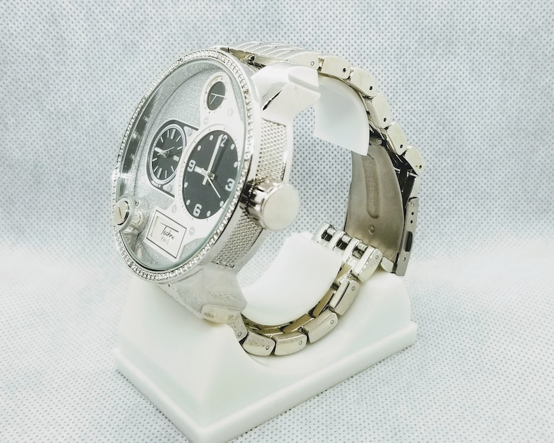 May include: A silver wristwatch with a multi-layered dial and a silver metal band. The watch is on a white display stand.