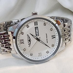 Op de afbeelding: Zilverkleurig polshorloge met een witte wijzerplaat, Romeinse cijfers en diamantaccenten. Het horloge toont de dag van de week en is waterbestendig. De band is een bijpassende zilveren schakelstijl.