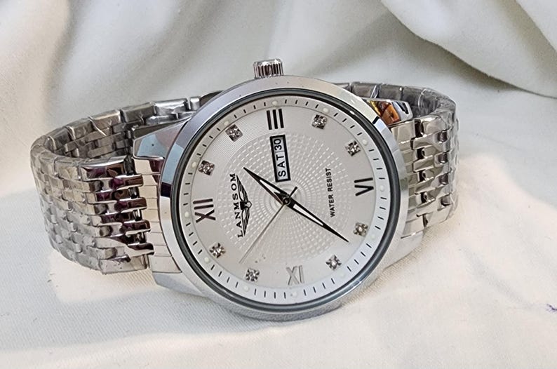 Op de afbeelding: Zilverkleurig polshorloge met een witte wijzerplaat, Romeinse cijfers en diamantaccenten. Het horloge toont de dag van de week en is waterbestendig. De band is een bijpassende zilveren schakelstijl.