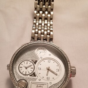May include: A silver Techno Pave watch with a white face and a silver band. The watch has a large, round face with a smaller, round face inside it. The watch has a silver band with a clasp.