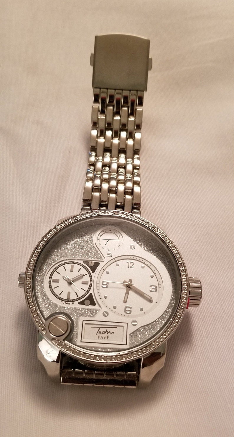 May include: A silver Techno Pave watch with a white face and a silver band. The watch has a large, round face with a smaller, round face inside it. The watch has a silver band with a clasp.